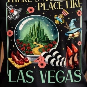 May include: A black t-shirt featuring a graphic print with the text "THERE'S NO PLACE LIKE LAS VEGAS". The design includes a green castle inside a snow globe, red poppies, a silver oil can, a blue hat, and legs in striped stockings with red shoes.