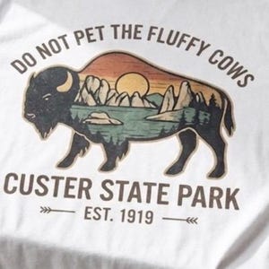 May include: A white t-shirt featuring a graphic of a bison silhouette filled with a landscape of mountains, trees, and a sun. The text reads DO NOT PET THE FLUFFY COWS, CUSTER STATE PARK, and EST. 1919 with small arrow icons on either side of the date.