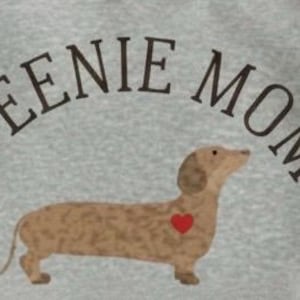 Puede incluir: Una sudadera gris claro con el texto "WEENIE MOM" en una fuente curva de color marrón oscuro sobre un perro salchicha marrón con un corazón rojo en su costado. El perro salchicha está mirando hacia la derecha.
