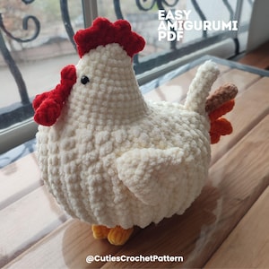 Może przedstawiać: Ręcznie robiony kurczak amigurumi z kremowej przędzy. Posiada czerwony grzebień i korale, z pomarańczowymi i brązowymi pi&oacute;rami ogona. Widoczny jest tekst "EASY AMIGURUMI PDF". Na dole obrazu znajduje się tekst "@CutiesCrochetPattern".