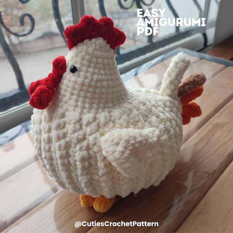 Może przedstawiać: Ręcznie robiony kurczak amigurumi z kremowej przędzy. Posiada czerwony grzebień i korale, z pomarańczowymi i brązowymi pi&oacute;rami ogona. Widoczny jest tekst "EASY AMIGURUMI PDF". Na dole obrazu znajduje się tekst "@CutiesCrochetPattern".