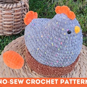 May include: A handmade crochet chicken in a nest. The chicken is gray with colorful flecks, orange comb and wattle, and a black eye. The nest is brown. An orange egg is next to the nest. The text "NO-SEW CROCHET PATTERN" is at the bottom.