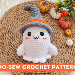 May include: A white crocheted ghost wearing a gray and orange witch hat. The ghost has black eyes and a small, pink mouth. The ghost is sitting on a woven straw mat with pumpkins and gourds around it. The text "NO-SEW CROCHET PATTERN" is at the bottom of the image.