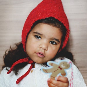 Puede incluir: Una niña pequeña con un gorro rojo de ganchillo y una bufanda a juego, sosteniendo un recorte de galleta de jengibre.