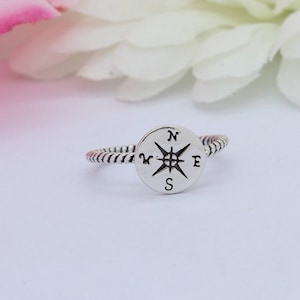 May include: A silver ring with a compass design. The compass features the letters N, S, E, and W, and a star in the center.