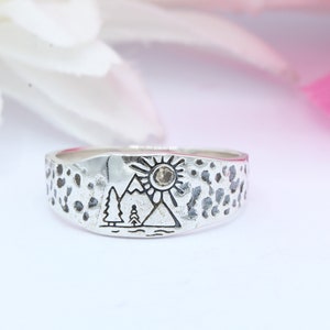 May include: A silver ring with a textured surface and a design of a mountain range with a sun and two trees. The sun has a small clear stone in the center.