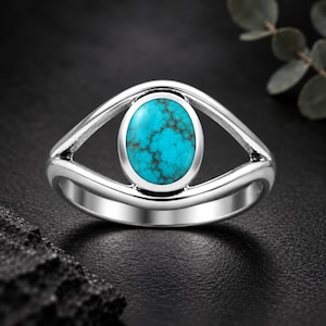 May include: A silver ring featuring a central oval-shaped turquoise gemstone with dark matrix veining. The band has a split design that curves around the stone, creating an open, eye-like frame around the polished blue-green cabochon.