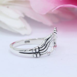 May include: A silver ring with two hands clasped together forming a heart shape. The ring is made of sterling silver and has a polished finish.