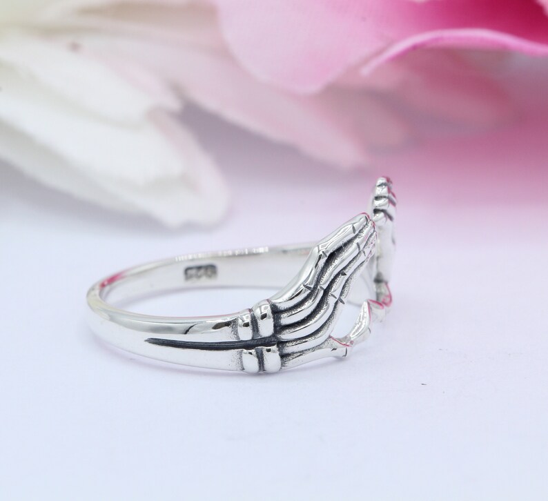 May include: A silver ring with two hands clasped together forming a heart shape. The ring is made of sterling silver and has a polished finish.