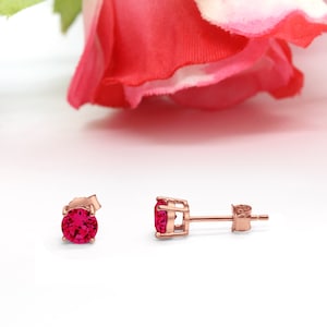 May include: A pair of rose gold stud earrings with a round, bright red ruby gemstone in each earring.