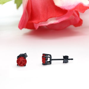 May include: A pair of black stud earrings with a red gemstone in the centre. The earrings are set in a black metal setting.