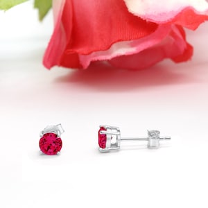 May include: A pair of stud earrings featuring round, vibrant red gemstones set in a silver metal. The earrings are displayed on a white surface with a blurred red rose in the background. The earrings are designed for pierced ears.