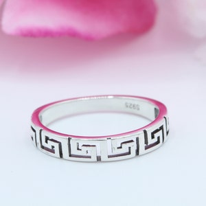 May include: A silver ring with a Greek key design. The ring has a polished finish and a repeating geometric pattern around the band. The ring is shown against a soft pink and white background.