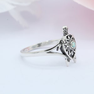 May include: A silver ring featuring a detailed turtle design with an opal gemstone in the center. The ring has a delicate, openwork design and a smooth band.