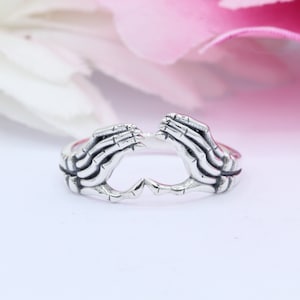 May include: A silver ring with two skeletal hands forming a heart shape.