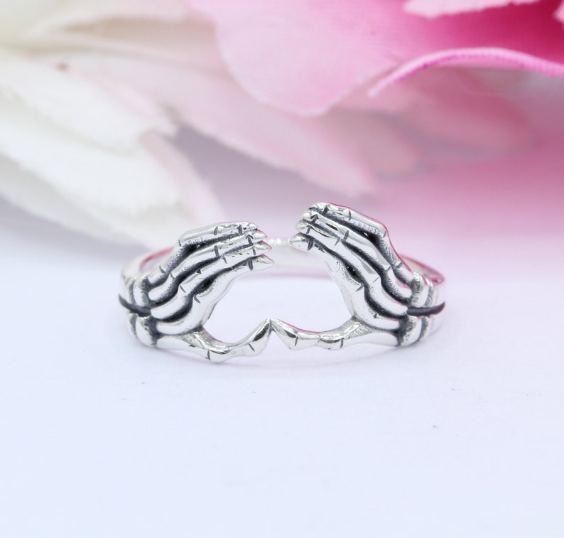 May include: A silver ring with two skeletal hands forming a heart shape.
