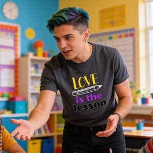 May include: A dark gray t-shirt with the words "LOVE is the lesson" in yellow and purple, with a pencil graphic. The person in the image is wearing black jeans and has colorful hair.