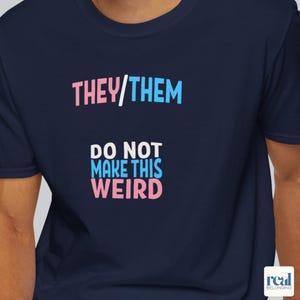 May include: A navy blue t-shirt featuring the words "THEY/THEM" in pink and blue, and "DO NOT MAKE THIS WEIRD" in the same colours. The shirt has a crew neck and short sleeves.
