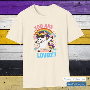 May include: Cream-colored t-shirt featuring a cartoon unicorn dabbing with sunglasses, a rainbow, and the text "YOU ARE LOVED!!!". The design includes stars and clouds. The shirt is shown in natural color.