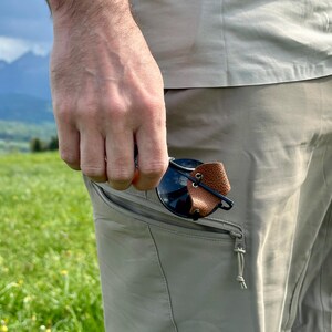 Peut inclure: Une personne portant un pantalon kaki avec une poche zippée. Une paire de lunettes de soleil avec une sangle en cuir marron est rangée dans la poche.
