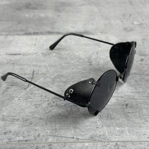 May include: Black round sunglasses with a leather side shield. The sunglasses have a metal frame and black lenses.