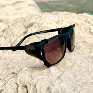 May include: Black wraparound sunglasses with a side shield and brown tinted lenses. The sunglasses are on a light brown rock surface.