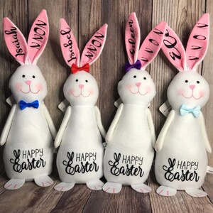May include: Four white plush Easter bunny decorations with pink ears. Each bunny has a different name on its ears: Isaiah, Isabel, Eileen, and El. Each bunny has a blue bow tie and the text "Happy Easter" on its belly.