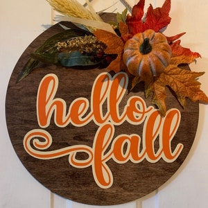 K&ouml;nnte beinhalten: Ein rundes Holzschild mit den Worten "hello fall" in orangefarbener Schrift mit wei&szlig;em Umriss. Das Schild ist mit Herbstlaub verziert, darunter ein kleiner orangefarbener K&uuml;rbis.