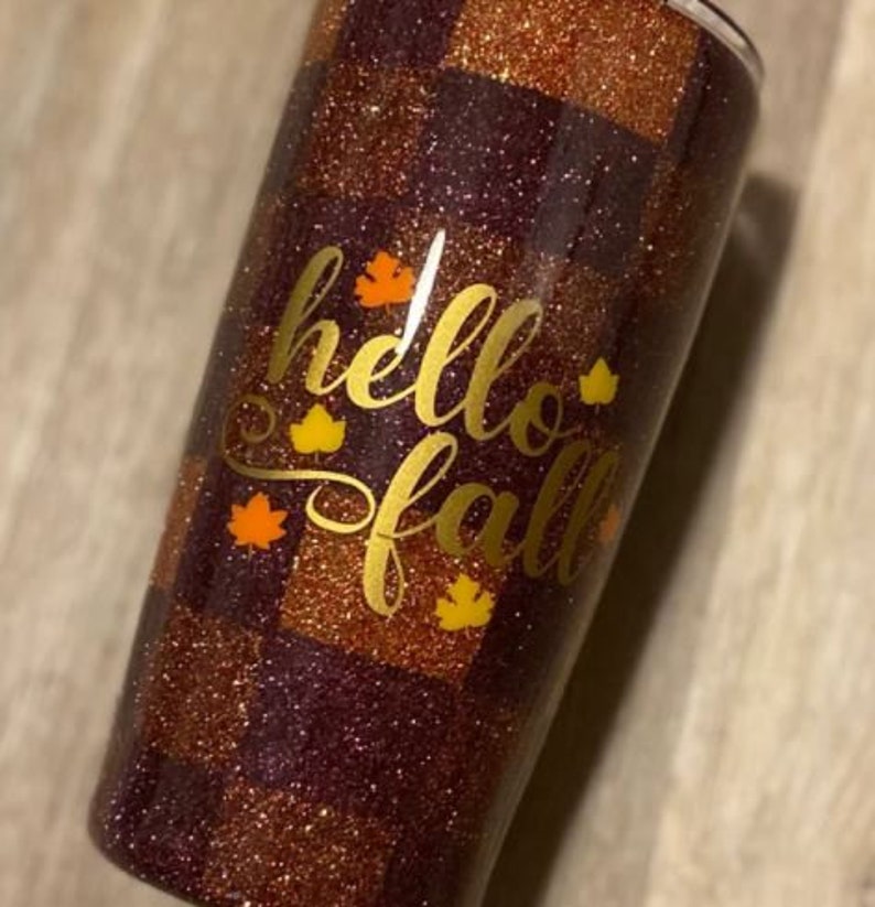 K&ouml;nnte beinhalten: Ein maronfarbener und goldfarbener Glitzerbecher mit einem karierten Muster. Der Becher tr&auml;gt die Aufschrift "hello fall" in goldfarbenem Glitzer mit kleinen goldfarbenen Glitzerblattakzenten.