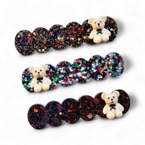 May include: Three glittery hair clips with a repeating oval design. Each clip features a small, white teddy bear with a black bow. The glitter is a mix of colors, including brown, red, and green, creating a sparkling effect.