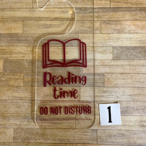 May include: Clear acrylic door hanger with a curved top and rectangular base. The design features a red open book icon, the words "Reading time" and "DO NOT DISTURB". A white square with the number "1" is in the lower right corner.