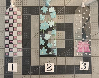 May include: Three decorative bookmarks with sheer ribbon ties. One features a black and white checkered pattern with pink bows. Another has blue flowers on a black background. The third has white hearts and a pink gift box.
