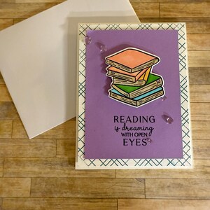 May include: A greeting card with a stack of books illustration on a purple background. The card reads "READING is dreaming WITH OPEN EYES." The card is decorated with a patterned border and comes with a white envelope.