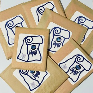 Puede incluir: Varios paquetes de papel marrón con etiquetas blancas que presentan un diseño de pergamino azul y la palabra "Oops!". Las etiquetas también incluyen un código QR y un pequeño gráfico. Los paquetes están sobre una superficie blanca.