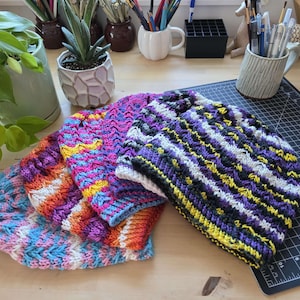 May include: Four colorful, hand-knitted beanies in shades of pink, purple, blue, orange, and yellow. Each hat features a textured, chevron-like pattern. The beanies are displayed on a wooden surface, suggesting a handmade craft item.