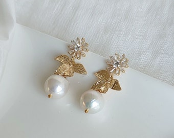 Puede incluir: Un par de pendientes de oro con una perla colgante y un pequeño diseño de flor con brillantes transparentes.