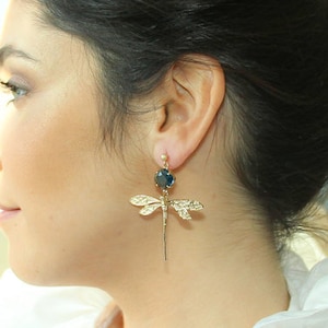 Può includere: Orecchini a forma di libellula dorati con una pietra preziosa blu scuro. Le ali della libellula sono dettagliate e gli orecchini pendono da una sottile catena d'oro.