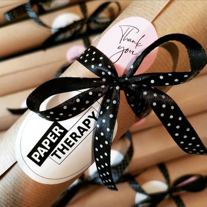 May include: A brown paper tube with a black and white polka dot ribbon tied around it. The tube has a white label with the text "PAPER THERAPY" on it. A pink circle sticker with the text "Thank you" is attached to the ribbon.