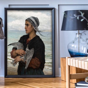 May include: A framed painting of a woman in a blue and white headscarf holding a seagull in her arms. The woman is standing on a beach with the ocean in the background.