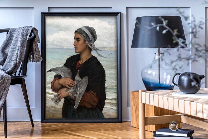 May include: A framed painting of a woman in a blue and white headscarf holding a seagull in her arms. The woman is standing on a beach with the ocean in the background.