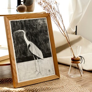 May include: A framed black and white illustration of a heron standing on a white surface. The frame is made of light brown wood and the illustration is set against a dark background.