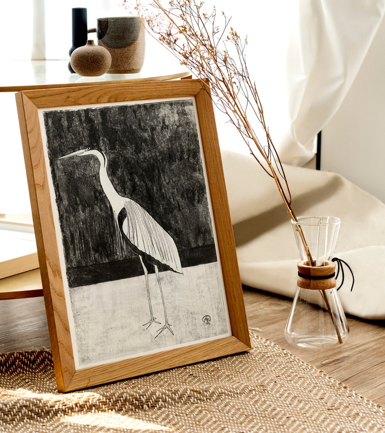 May include: A framed black and white illustration of a heron standing on a white surface. The frame is made of light brown wood and the illustration is set against a dark background.