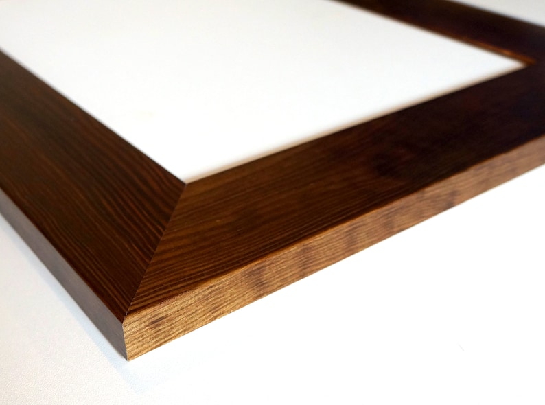 Puede incluir: Primer plano de un marco de fotos rectangular de madera con acabado marr&oacute;n oscuro. El marco tiene una textura de veta de madera natural y un interior blanco. El marco est&aacute; en &aacute;ngulo, mostrando una esquina y parte del lado.