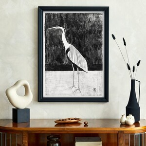 May include: A black and white framed print of a heron standing on a white background. The print is hanging on a wall above a wooden console table with a white ceramic sculpture, a vase with dried flowers, and a wooden bowl with nuts.