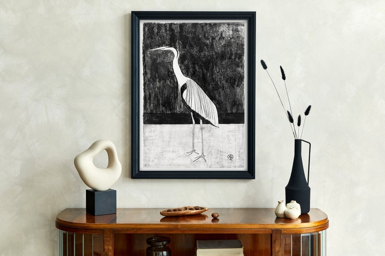 May include: A black and white framed print of a heron standing on a white background. The print is hanging on a wall above a wooden console table with a white ceramic sculpture, a vase with dried flowers, and a wooden bowl with nuts.