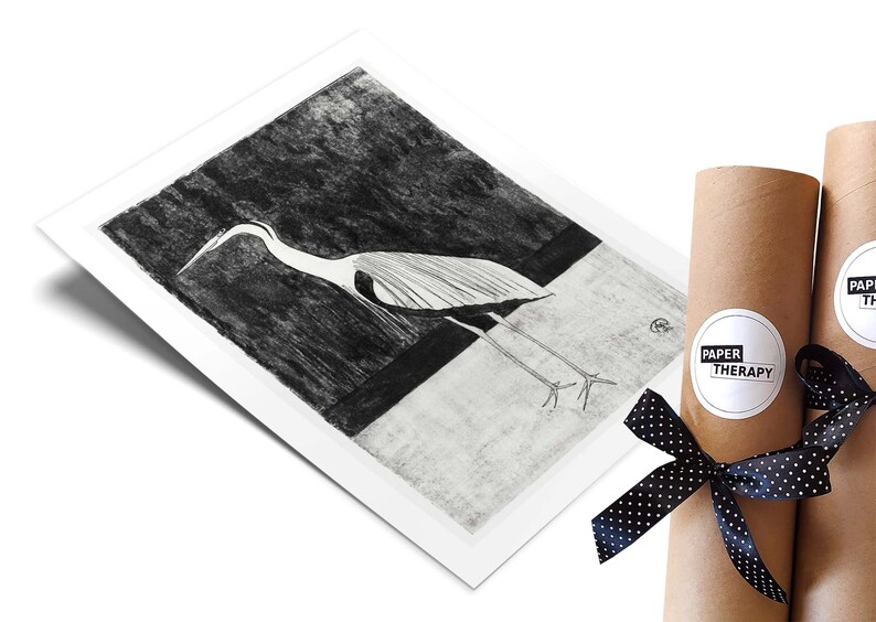 May include: Black and white illustration of a heron standing on a white surface with a dark background. The illustration is printed on a white paper and rolled up in a brown tube with a black and white polka dot ribbon and a white sticker with the text "PAPER THERAPY".