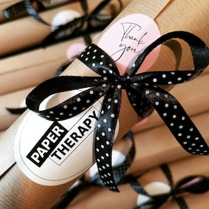 May include: A brown paper tube with a black and white polka dot ribbon tied around it. The tube has a white sticker with the text "PAPER THERAPY" on it. A pink sticker with the text "Thank you" is also on the tube.