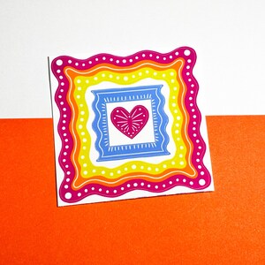 May include: A colorful sticker featuring a heart design within layered frames. The sticker has a white background with a vibrant orange and white border. The heart is pink, and the frames are blue, yellow, and pink.