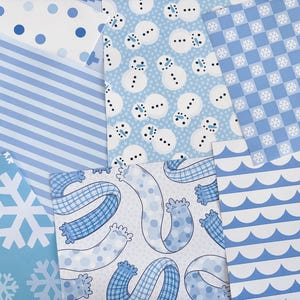 Könnte beinhalten: Eine Auswahl an hellblauen und weißen Papieren mit Mustern. Designs umfassen Schneeflocken, Schneemänner, Streifen, Punkte und Wellenlinien. Die Papiere sind in einer geschichteten, überlappenden Anordnung angeordnet und zeigen die Vielfalt der winterlichen Muster.