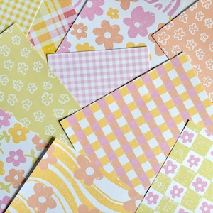 Pode incluir: Uma coleção de folhas de papel estampadas com vários desenhos e cores. Os padrões incluem xadrez, floral e listras em tons de amarelo, rosa e laranja. As folhas estão dispostas de forma sobreposta.